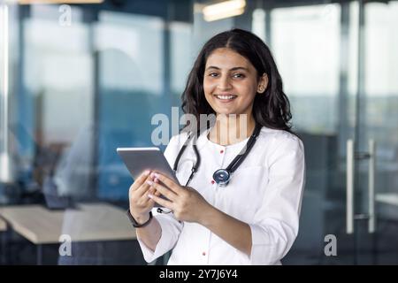 Giovane donna medico con fiducia utilizzando tablet con stetoscopio in una moderna struttura sanitaria. L'abbigliamento professionale e il sorriso cordiale riflettono la dedizione alla cura e alla tecnologia dei pazienti. Foto Stock