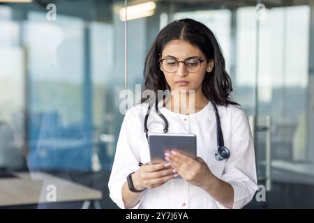 Giovane medico donna sicura di sé utilizza lo smartphone per la ricerca medica e la comunicazione in ufficio moderno. Indossa un cappotto bianco e uno stetoscopio che trasmettono professionalità e attenzione. Foto Stock