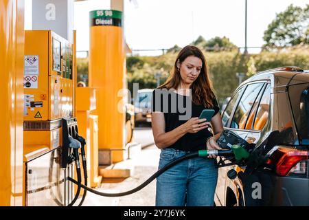 Donna che usa lo smartphone mentre rifornisce l'auto al distributore di benzina Foto Stock