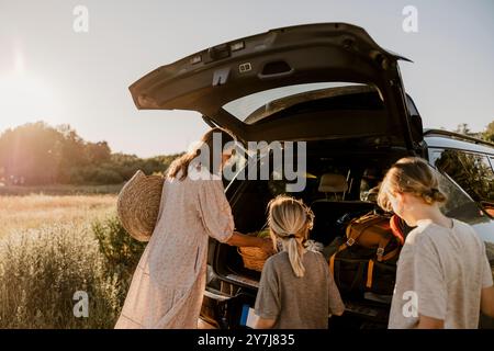 Mamma che scarica il cestino dal bagagliaio dell'auto con i ragazzi sul prato durante la giornata di sole Foto Stock