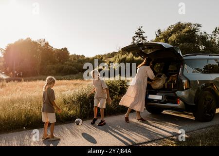 Madre vicino al bagagliaio della macchina con ragazzi che giocano con la palla da calcio sul vialetto del prato nelle giornate di sole Foto Stock