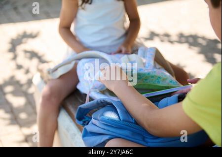 Due bambini seduti all'esterno, che esplorano il contenuto dei loro zaini in una giornata di sole. La scena cattura un momento di curiosità e amicizia. Foto Stock