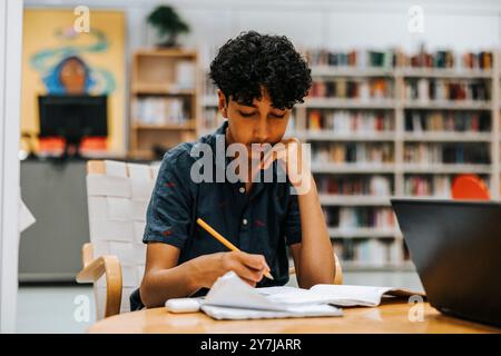 Adolescente concentrato che studia con la mano sul mento mentre siede vicino a un tavolo in biblioteca alla scuola media Foto Stock