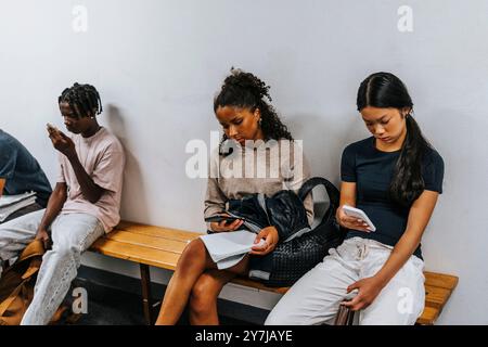 Studenti di sesso maschile e femminile che usano gli smartphone mentre sono seduti sulla panchina vicino al muro a scuola Foto Stock