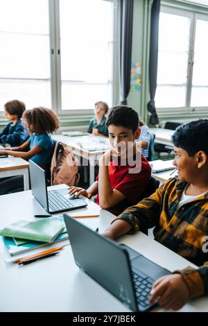 Studenti che studiano con un computer portatile mentre sono seduti in classe alla scuola elementare Foto Stock