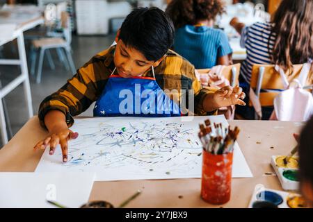 Ragazzo concentrato che dipinge le dita su carta durante la lezione d'arte alla scuola elementare Foto Stock