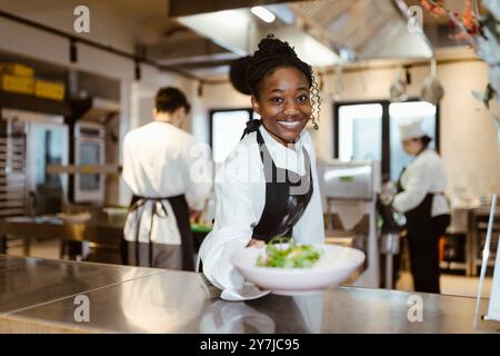 Ritratto di un giovane chef sorridente che passa davanti a una ciotola per insalate nella cucina commerciale del ristorante Foto Stock