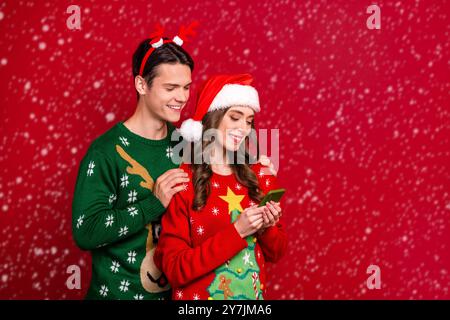 Ritratto di due persone abbracciare utilizzare telefono eshop media app santa Headwear corna di renna isolato su sfondo di colore rosso Foto Stock