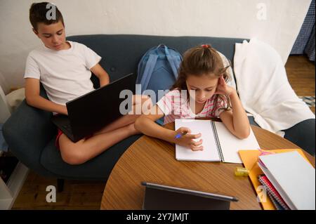 Due bambini si dedicavano allo studio a casa, utilizzando notebook e notebook. Un ragazzo si concentra sul suo notebook mentre una ragazza scrive su un notebook. Foto Stock