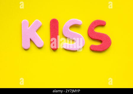 La parola bacio dalle lettere rosse e rosa dell'alfabeto su sfondo giallo con spazio di copia. Vista dall'alto, disposizione piatta. Foto Stock