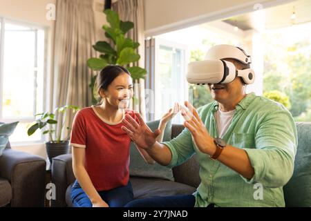 Utilizzando il visore VR, il padre interagisce mentre la figlia sorride in salotto Foto Stock