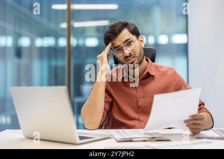 L'uomo in ufficio stressato che tiene un documento che mostra confusione e preoccupazione, seduto alla scrivania con un laptop aperto. Professionisti aziendali che soffrono di stress durante la gestione di documenti cartacei nei moderni ambienti di lavoro. Foto Stock