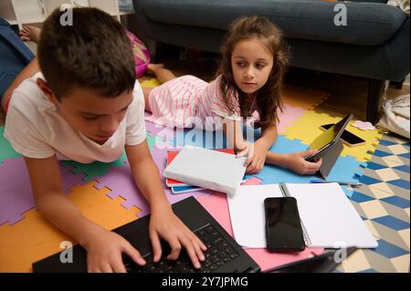 Due bambini si dedicano all'apprendimento online a casa utilizzando un notebook e un tablet, circondati da materiali didattici colorati. Foto Stock