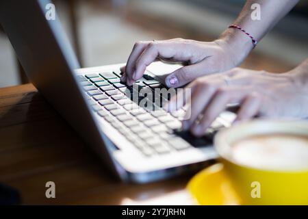 Un primo piano di mani che digitano sulla tastiera di un notebook, con una tazza di caffè in primo piano. L'ambiente sembra accogliente e invitante, con una luce calda e. Foto Stock