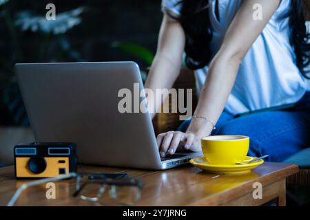 Una donna che scrive su un notebook in un ambiente accogliente. Una tazza di caffè gialla si trova su un piattino accanto al laptop e vengono posizionati una fotocamera vintage e degli occhiali Foto Stock