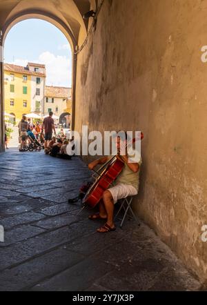 Un musicista suona all'ingresso di Piazza dell'Anfiteatro, una piazza pubblica nel quadrante nord-est della città fortificata di Lucca, in Toscana. Foto Stock