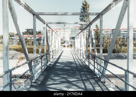Un'immagine grandangolare di un ponte pedonale in metallo con una struttura geometrica di travi e ringhiere, che si estende su un canale asciutto verso un'area residenziale Foto Stock