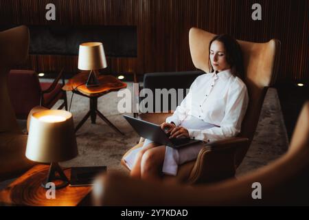 Una donna caucasica con una camicetta bianca formale e una gonna viola chiaro si siede rilassata in una poltrona con gli occhi chiusi, utilizzando un laptop in grembo in una mod Foto Stock
