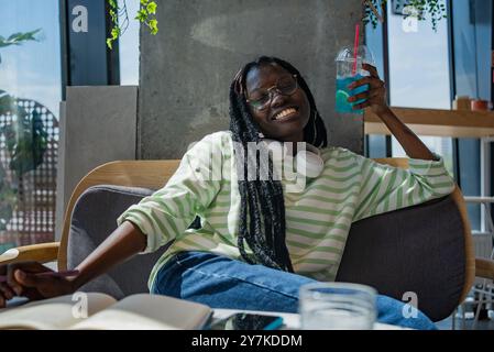 La donna afroamericana si rilassa con una bevanda blu e le cuffie, sorridendo in un accogliente ambiente da caffè, godendosi il suo tempo di riposo. Foto Stock