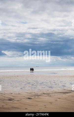Una coppia cammina mano nella mano verso il mare con la bassa marea con una nave sullo sfondo sotto un cielo nuvoloso con spazio per copiare a Cambois, Northumberland, Regno Unito Foto Stock