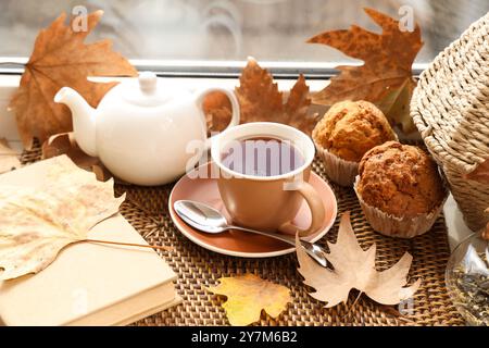 Teiera con tazza di bevanda calda, gustosi cupcake, libro e foglie autunnali sul davanzale in camera Foto Stock