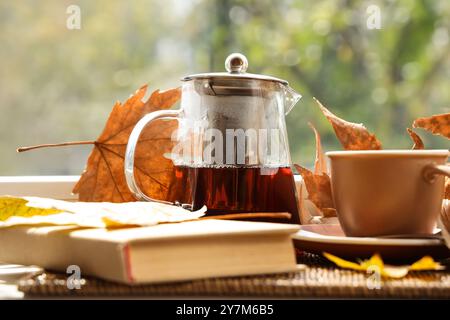 Teiera con tazza di bevanda calda, libro e foglie autunnali sul davanzale Foto Stock