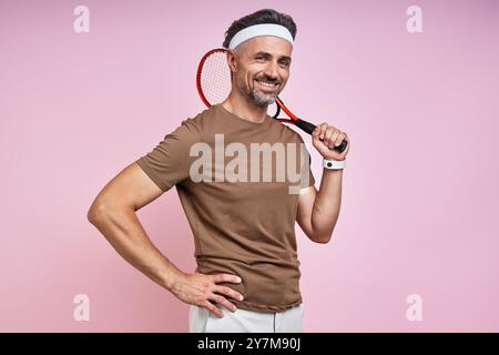 Allegro uomo maturo che porta una racchetta da tennis sulla spalla mentre è in piedi su sfondo rosa Foto Stock