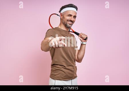 Uomo maturo felice che porta racchetta da tennis e telecamera puntando mentre è in piedi su sfondo rosa Foto Stock