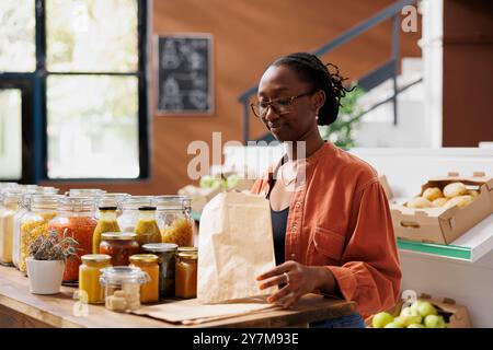 Donna nera al banco di legno che tiene in mano un sacchetto di carta per mettere prodotti alimentari biologici freschi al minimarket. Cliente afroamericano che afferra un piccolo sacco marrone per gli articoli appena raccolti presso il negozio locale. Foto Stock