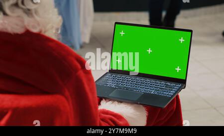 Il manager di Natale adorna il centro commerciale vestito da Babbo Natale che tiene un notebook con schermo verde, guarda il sito Web di e-commerce del negozio di abbigliamento e inserisce offerte promozionali per le vacanze Foto Stock