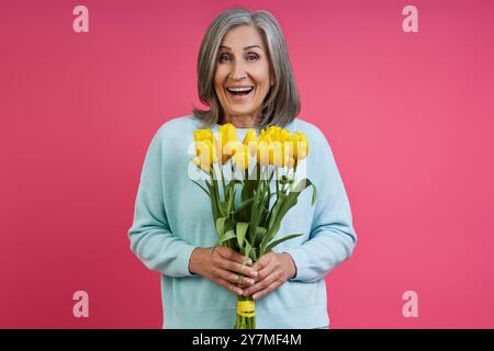 Una donna anziana sorpresa che tiene in mano un mucchio di tulipani mentre è in piedi su sfondo rosa Foto Stock