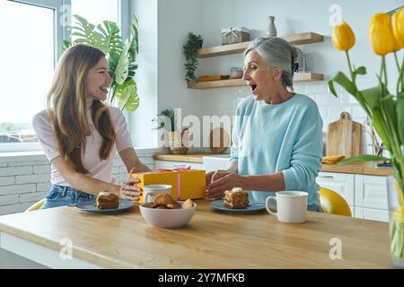 Madre anziana felice che riceve una confezione regalo dalla figlia adulta mentre entrambe sono sedute in cucina Foto Stock