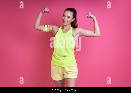 Donna felice con abbigliamento sportivo che indossa la fascia capitano e mostra bicipiti su sfondo rosa Foto Stock