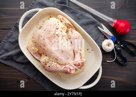 Pollo crudo condito in un piatto bianco da forno: Pollo intero con le gambe legate ricoperto di burro fuso, sale marino e pepe nero in una teglia da forno Foto Stock