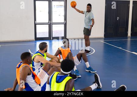 Allenatore di pallacanestro che tiene la palla e istruisce i giocatori della squadra seduti sul campo Foto Stock