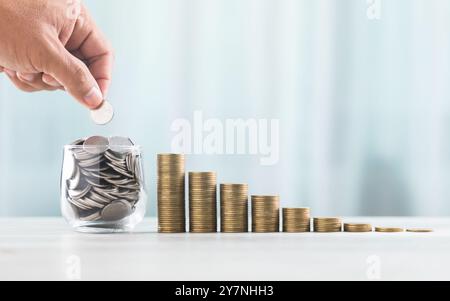 La mano getta le monete in un vaso di vetro, con pile di monete disposte dal più piccolo al più grande sul tavolo. Lo sfondo bianco e luminoso enfatizza Foto Stock