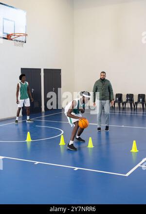 Dribbling di pallacanestro intorno ai coni, giocatore che pratica abilità con allenatore in palestra Foto Stock