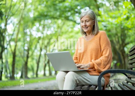 Donna anziana seduta sulla panchina del parco con un laptop, sorridente mentre ama la natura. Rappresenta il tempo libero all'aperto, il lavoro online e lo stile di vita digitale per gli adulti più anziani. Foto Stock