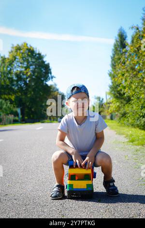 Un bambino sta guidando il suo luminoso e colorato camion giocattolo sulla strada sotto un cielo blu cristallino in campagna Foto Stock