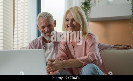 Maturo anziano di mezza età caucasica coppia sorridente donna allegra uomo ridere insieme vecchia famiglia guardare computer portatile schermo che parla a casa divano Foto Stock