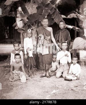 Fotografia d'epoca del Siam (Thailandia): Monaci e loro assistenti. Di John Thomson, 1865. Due scene diverse su un solo negativo. Sulla metà sinistra, un giovane monaco è seduto su una sedia al centro, mentre altri ragazzi sono seduti a terra su entrambi i lati. Sulla destra mezzo monaco anziano sta al centro, con ragazzi seduti, inginocchiati e in piedi a terra intorno a lui. Quattro di loro si prendono cura dei parapernali del monaco: Un tappetino di corsa, un ventilatore, una bara e una teiera. Foto Stock