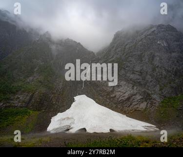 Grotta di ghiaccio annidata alla base di una montagna nebbiosa con una cascata che scorre giù scogliere rocciose, circondata da nebbia e nuvole in una remota natura selvaggia. Foto Stock