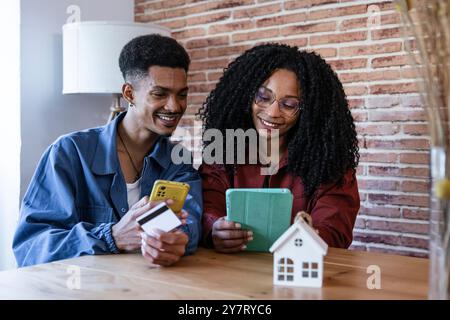 Felice coppia diversificata che acquista investimenti online e immobili finanziari a casa. Marito e moglie con carta di credito e smartphone alla ricerca di un nuovo Foto Stock