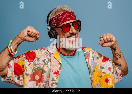 Uomo anziano felice con camicia e cuffie funky che ascolta la musica e balla su sfondo blu Foto Stock