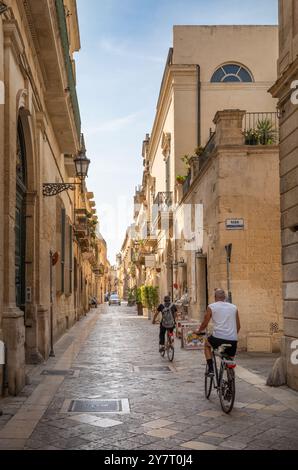 Vista posteriore di due persone in bicicletta lungo una stradina fiancheggiata da edifici in pietra nell'antica città di Lecce, Puglia, Italia. Foto Stock