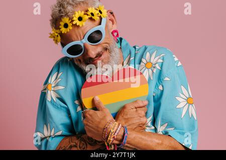 Maturo gay con corona floreale che abbraccia la bandiera arcobaleno a forma di cuore su sfondo rosa Foto Stock