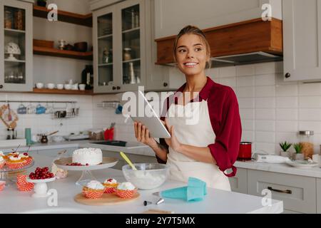 Una bellissima giovane pasticciera che usa un tablet digitale mentre prepara torte e muffin in cucina Foto Stock