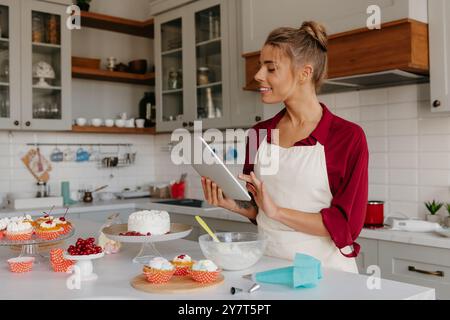 Pasticceria femminile sicura di sé che utilizza una tavoletta digitale per preparare torte e muffin in cucina Foto Stock