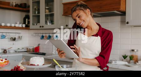 Felice giovane pasticciera che discute l'ordine mentre parla al telefono con il cliente in cucina Foto Stock
