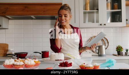 Una pasticciera sicura di sé che discute i dettagli dell'ordine mentre parla al telefono con il cliente in cucina Foto Stock